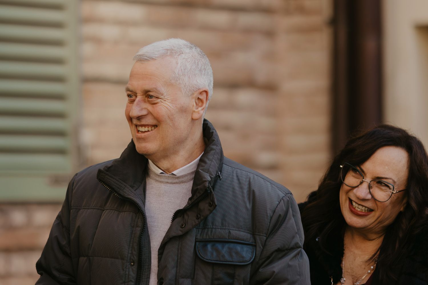 Two smiling middle-aged adults, a man with gray hair and a woman with dark hair and glasses, outdoors near a brick wall.