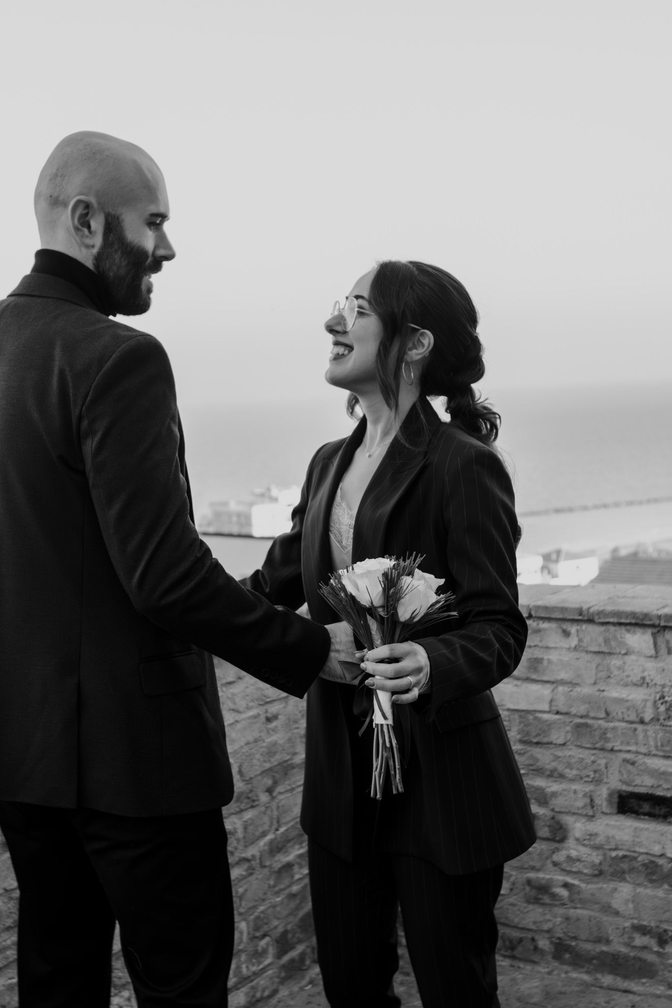 Black and white photo of a smiling couple holding flowers, standing on a stone terrace with ocean views.