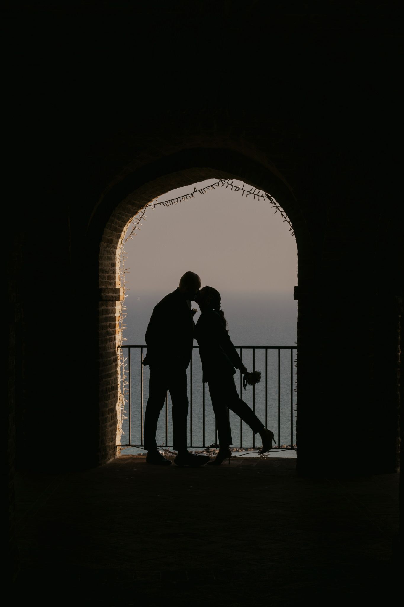 Romantic silhouette of a couple kissing under a brick archway at dusk, with a railing and twilight sky in the background.