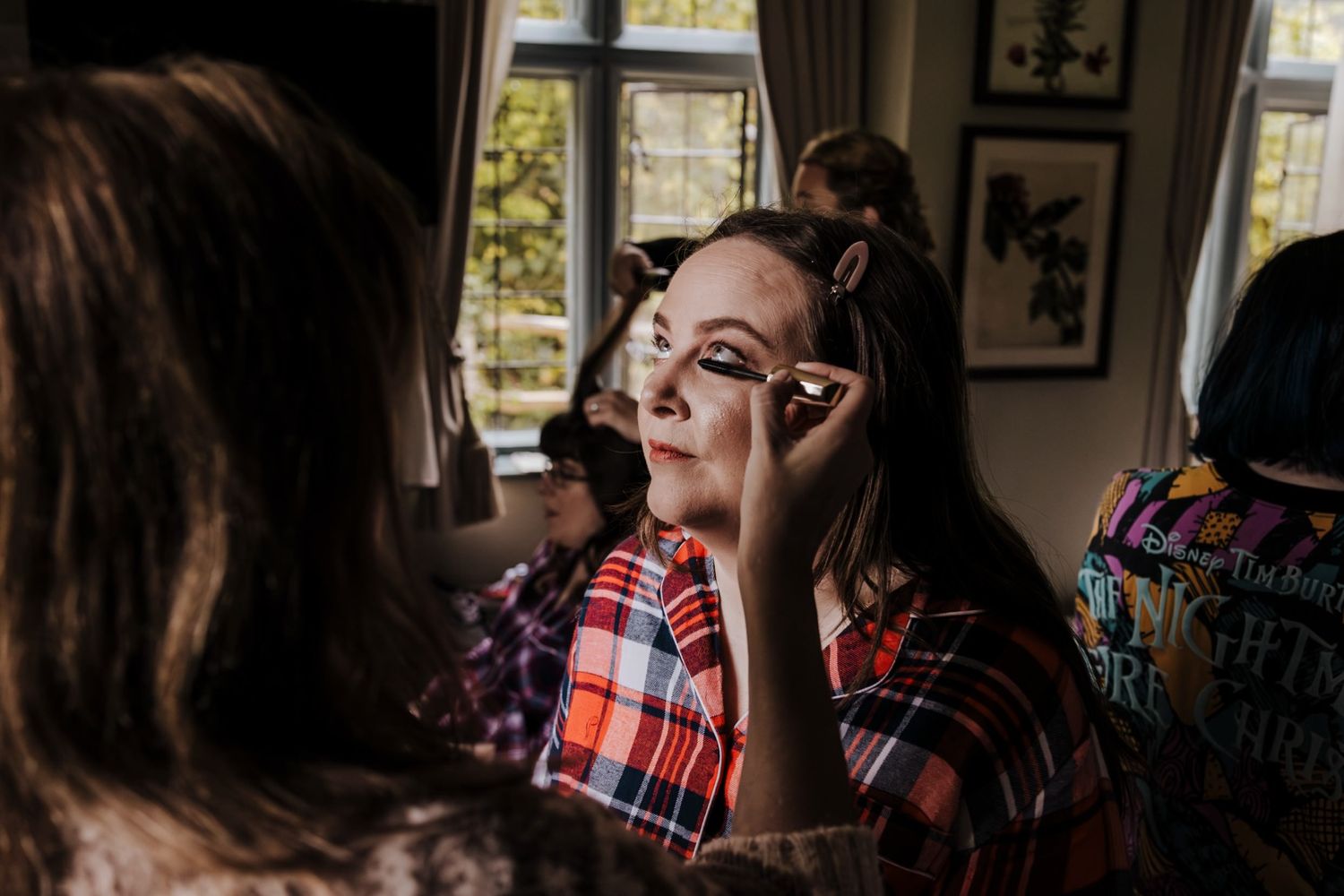 Bridesmaid getting makeup applied while sitting in a plaid shirt during wedding preparations indoors.
