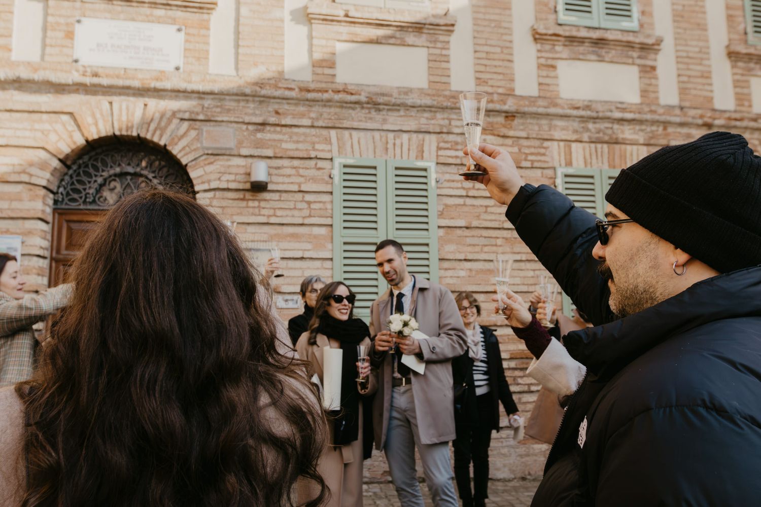 Group of people celebrating outdoors near a historic brick building with green shutters, raising glasses in a toast.