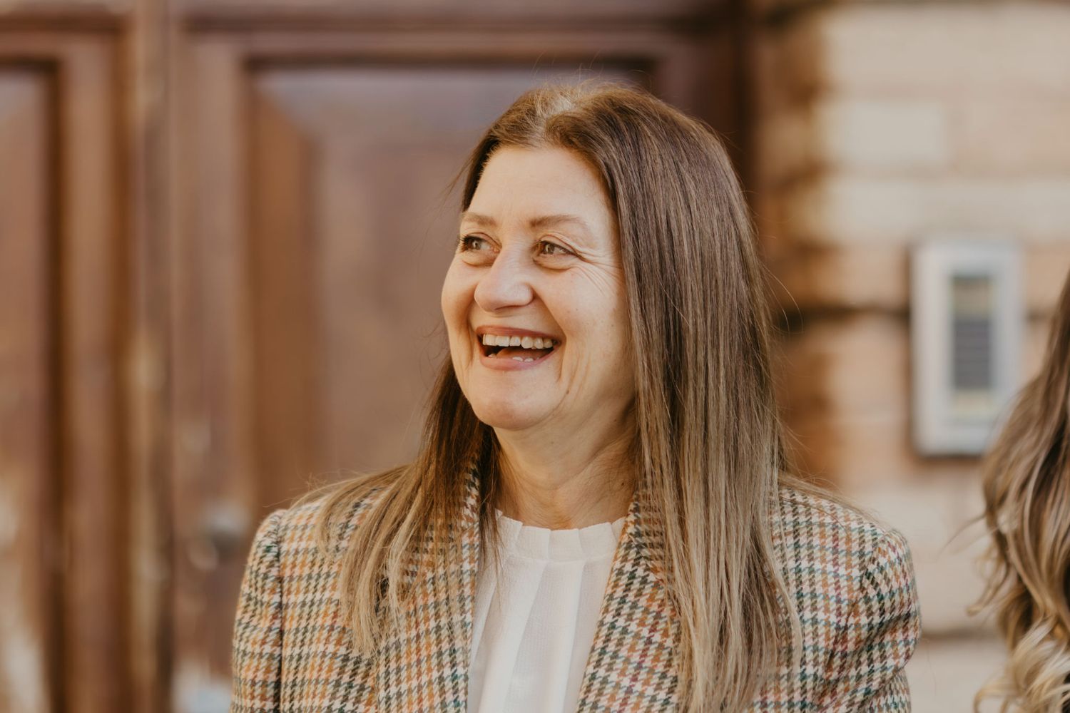 A woman with long hair laughs joyfully outdoors, wearing a plaid blazer over a white top.