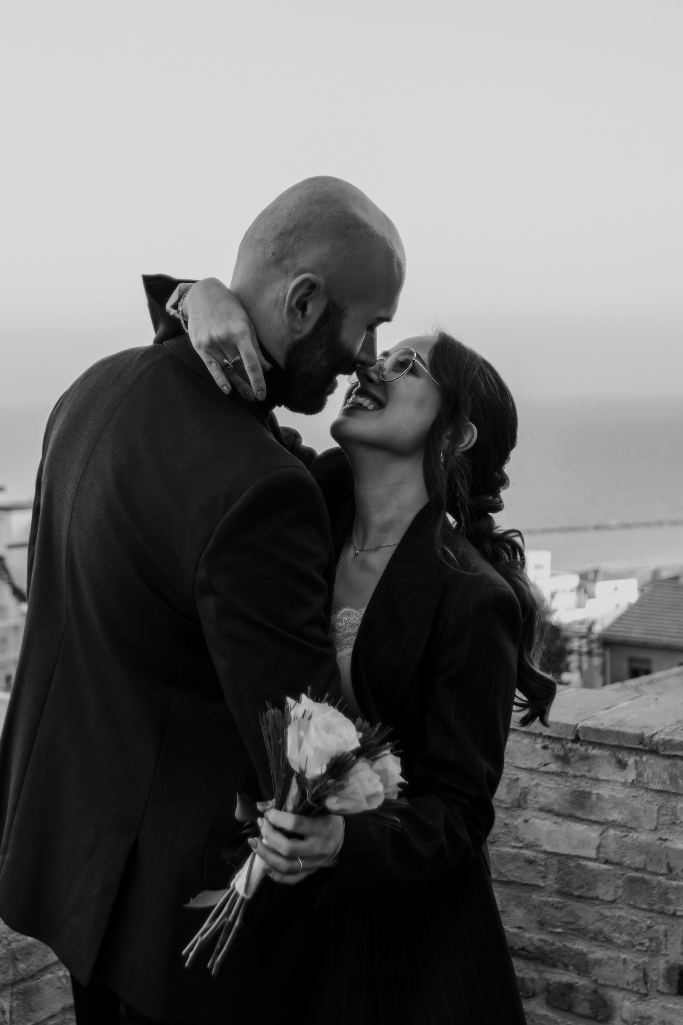 Black and white photo of a couple sharing a romantic moment, woman holding a bouquet, with a coastal town in the background.