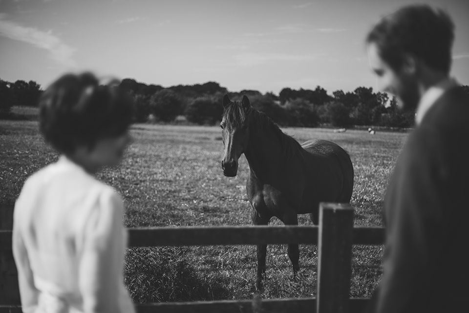 Black and white photo of a couple watching a horse standing by a wooden fence in a open field.