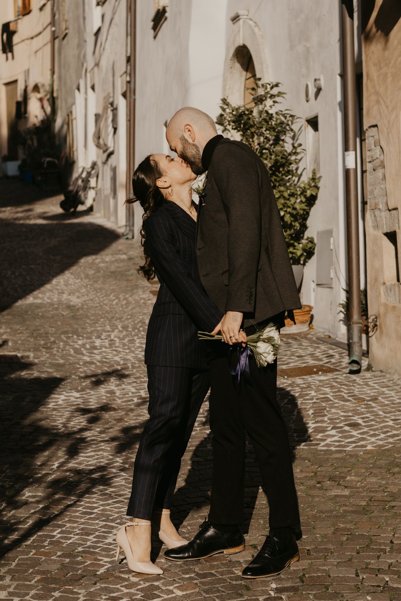 Couple kissing on cobblestone street in dark formal attire, holding white flowers, with sunlit European buildings behind them.