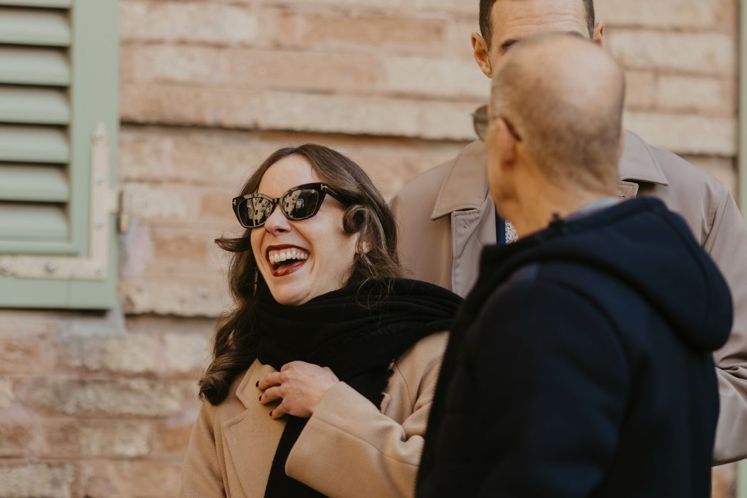 Woman in sunglasses and black scarf laughing joyfully with friends outdoors near a brick wall.