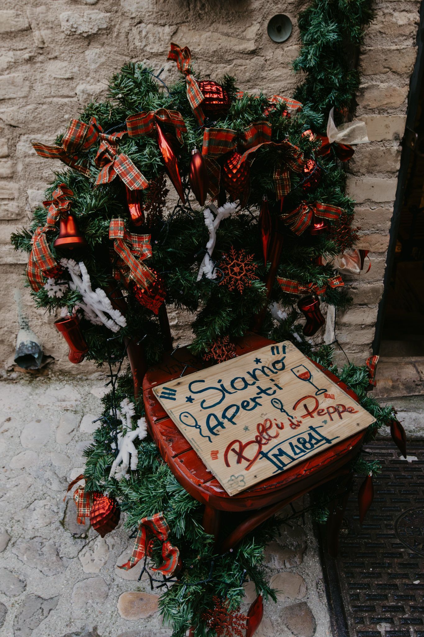 A festively decorated red chair with Christmas garland, ribbons, and a handwritten Italian sign outside a restaurant.