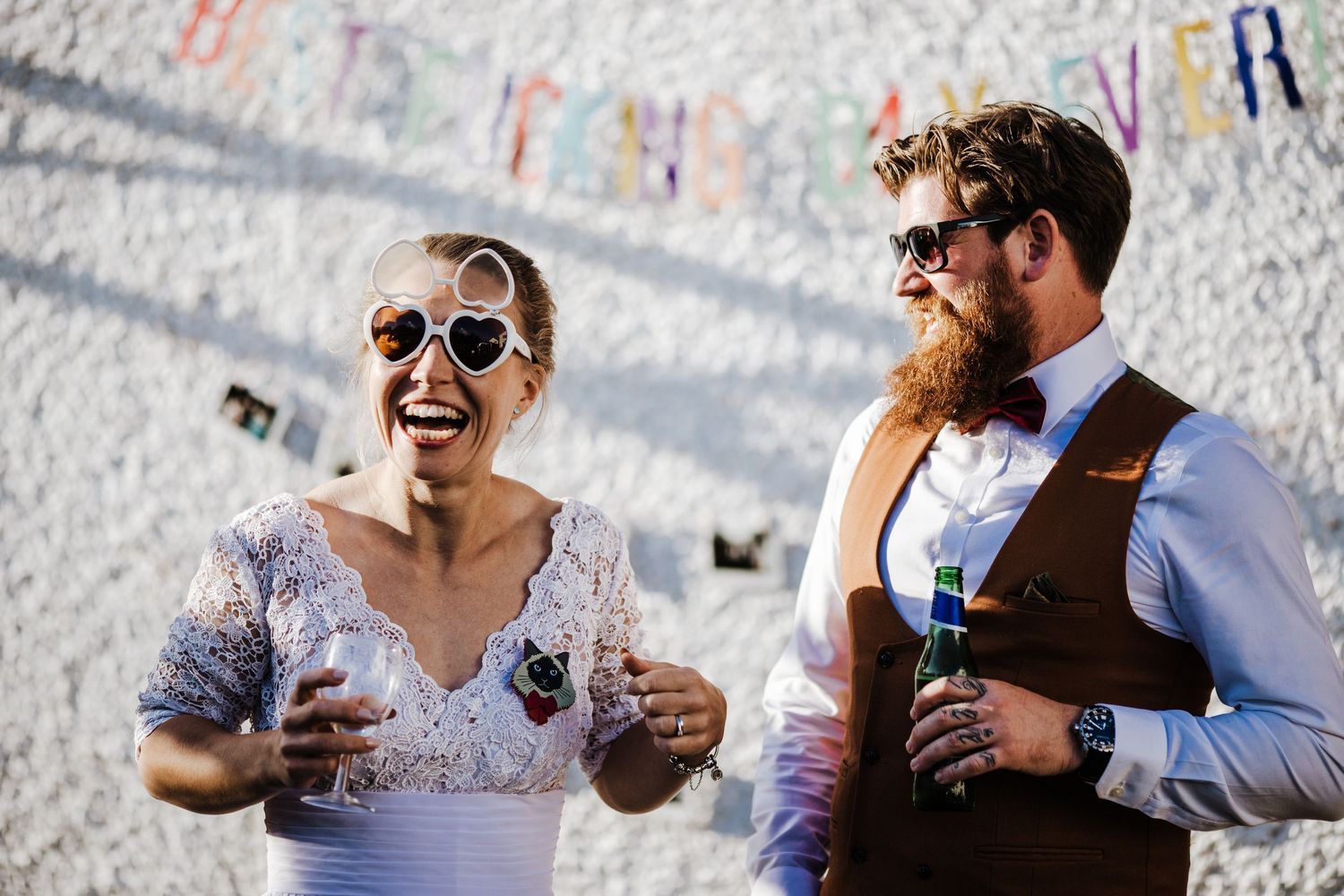 A couple in sunglasses share drinks and laughter at their wedding, outdoors on a sunny day.  - Glastonbury wedding photographer