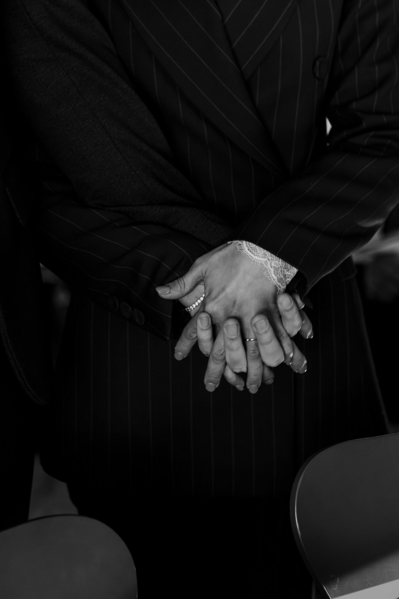 Black and white photo of clasped hands wearing rings, resting on lap in pinstripe suit jacket.