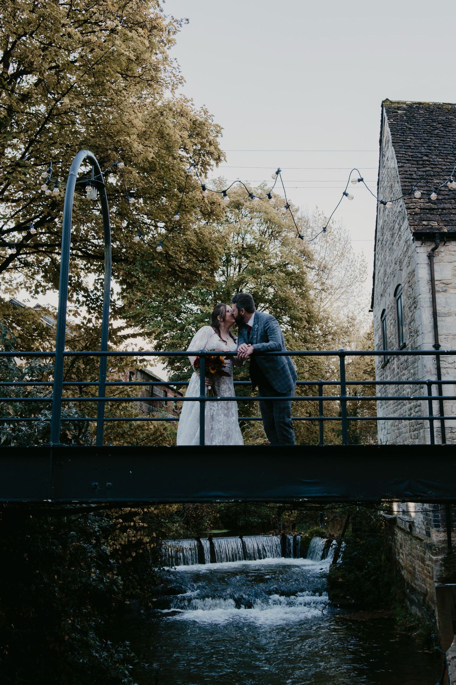Bride and groom share a romantic kiss on a bridge over a waterfall, surrounded by autumn trees and string lights.