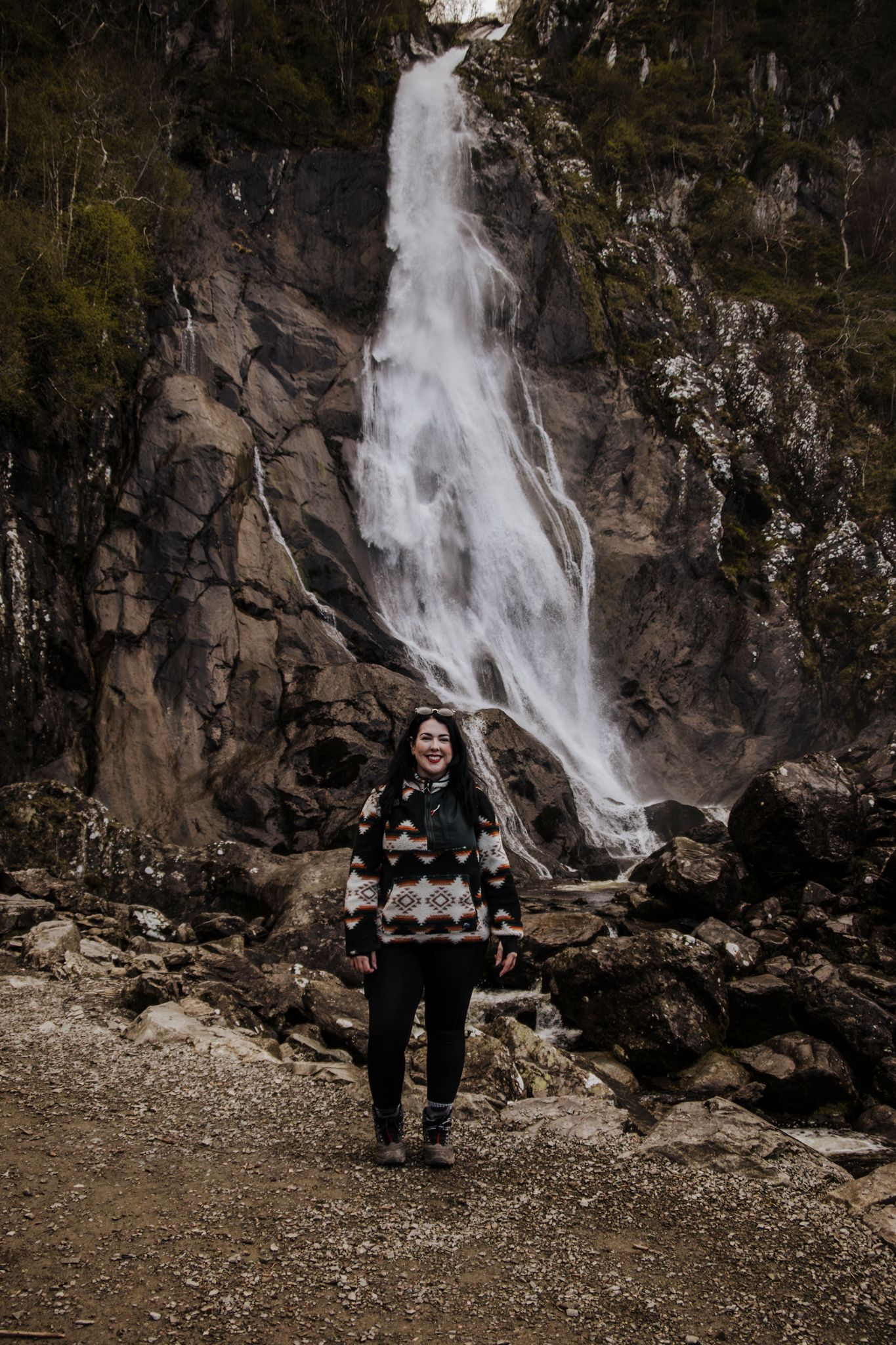 Woman in patterned sweater smiling in front of a tall cascading waterfall on a rocky mountain trail.