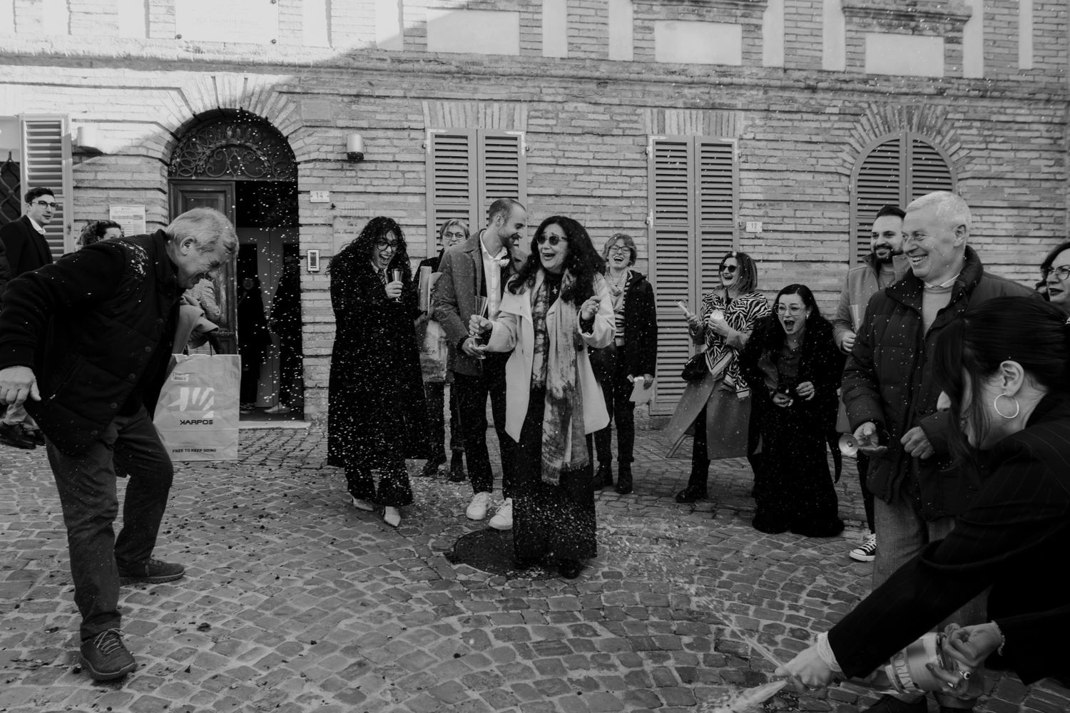 Black and white photo of a joyful group of people celebrating outdoors on a cobblestone street in Italy.