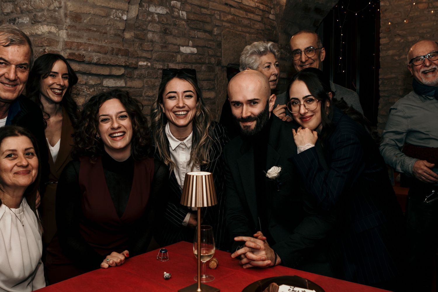 Group of people smiling together at a wedding reception in a rustic brick interior.