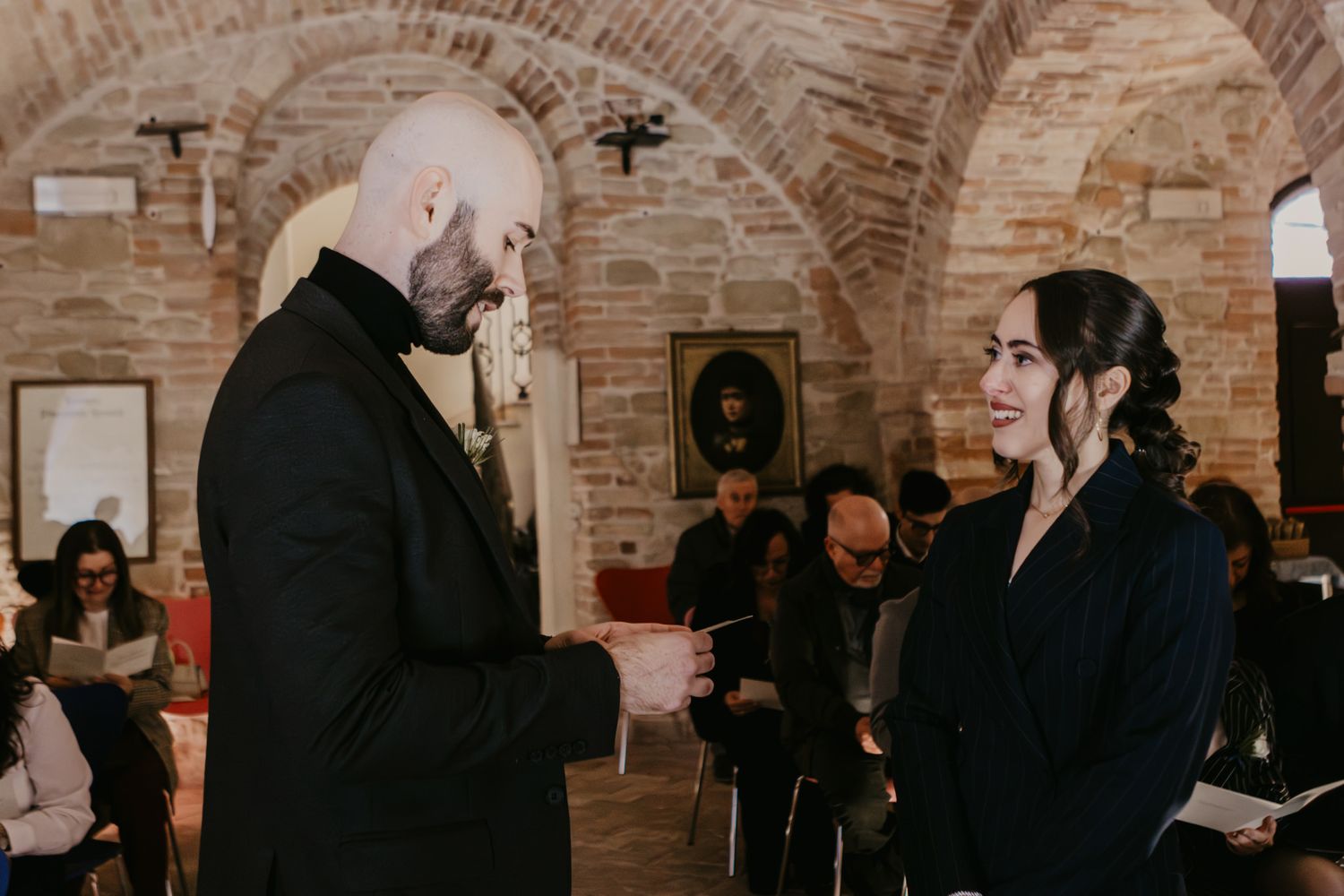 Couple in black attire exchanging vows in a rustic brick venue during an intimate wedding ceremony.
