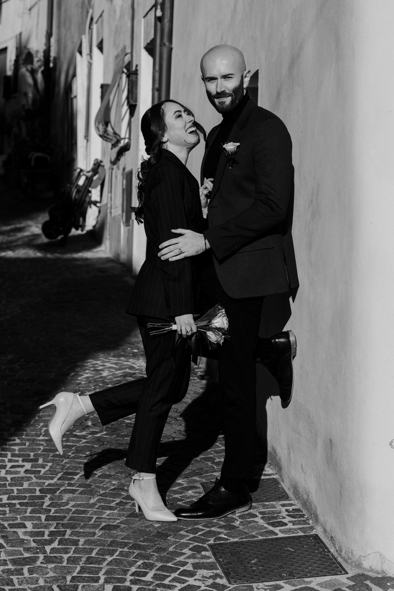 Black and white photo of a laughing couple embracing in a narrow cobblestone alley, dressed in elegant black attire.