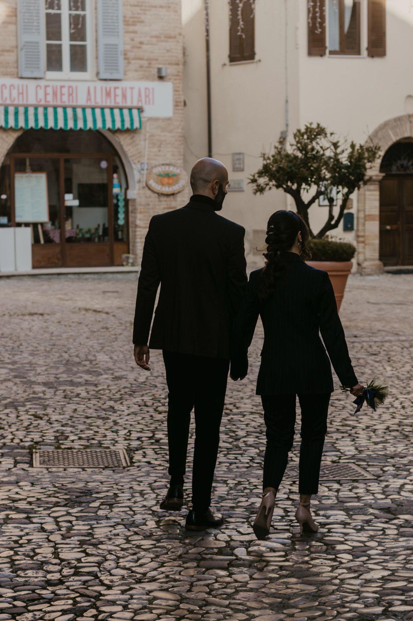 Couple in black suits walking hand-in-hand on cobblestone street in Italian village square.
