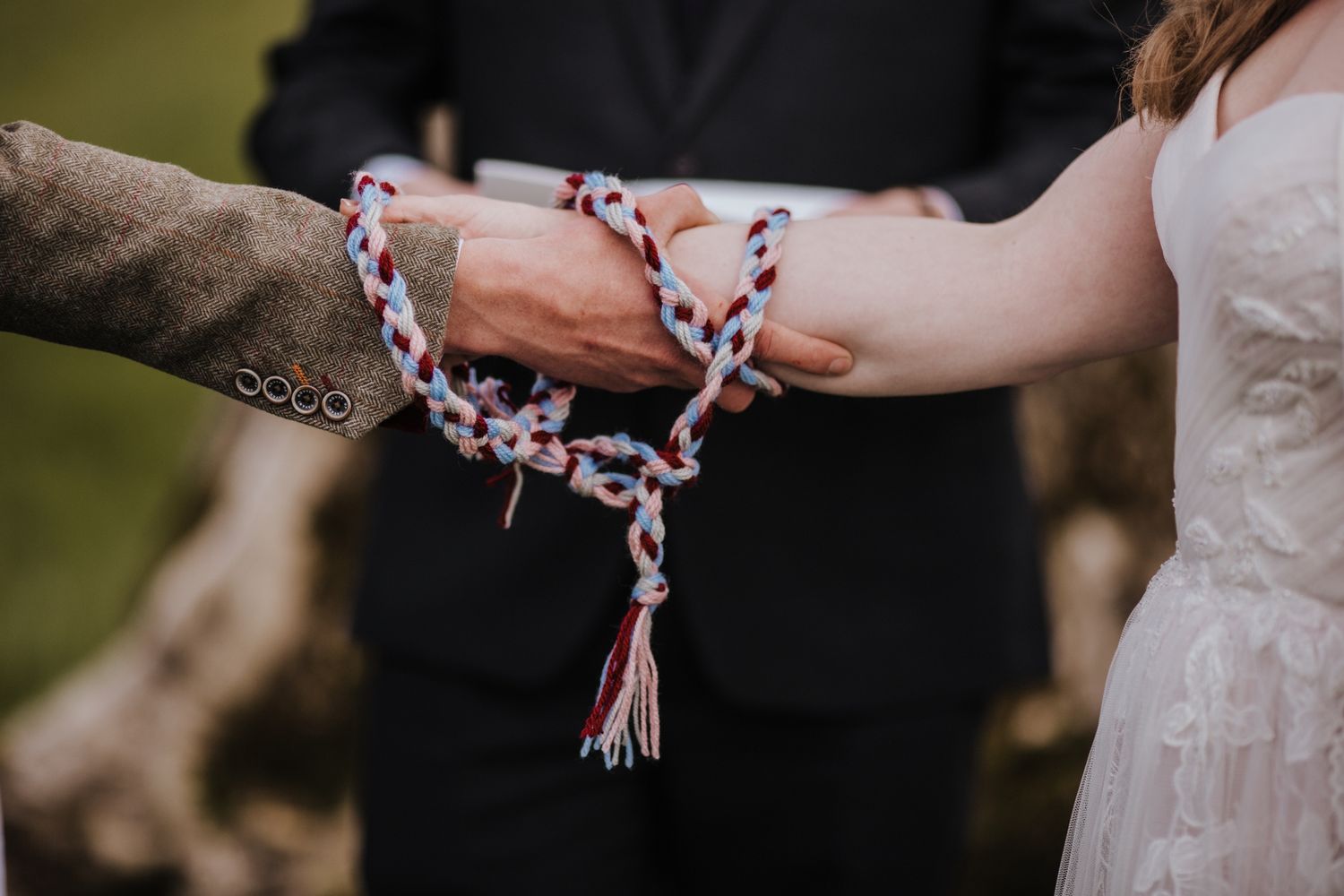 Couple's hands bound together with red, white, and blue braided cord during a handfasting wedding ceremony outdoors.