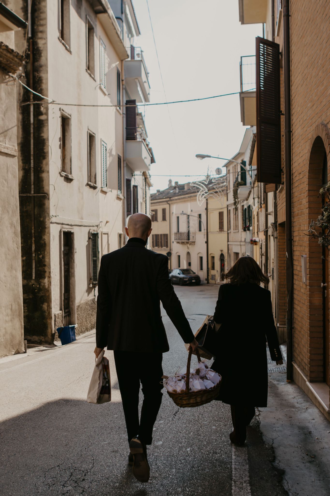 Couple walking hand in hand through a narrow Italian street, carrying shopping bags and a basket.
