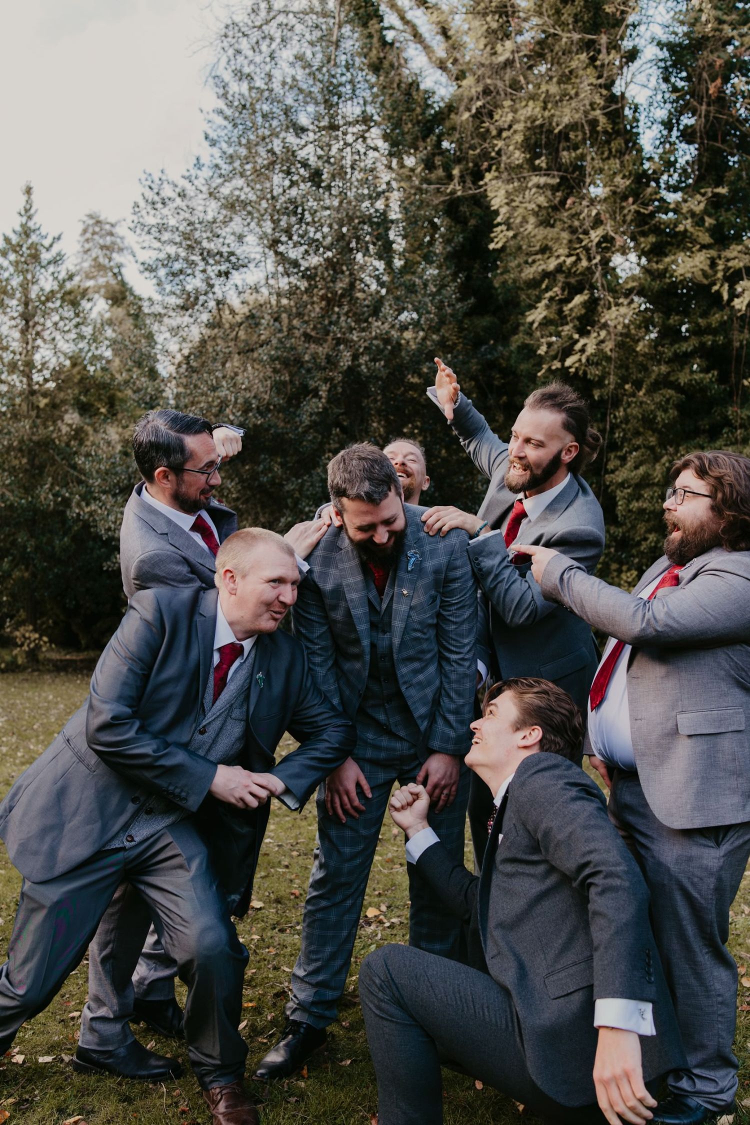 Groom surrounded by groomsmen in grey suits and red ties celebrating outdoors near tall evergreen trees.