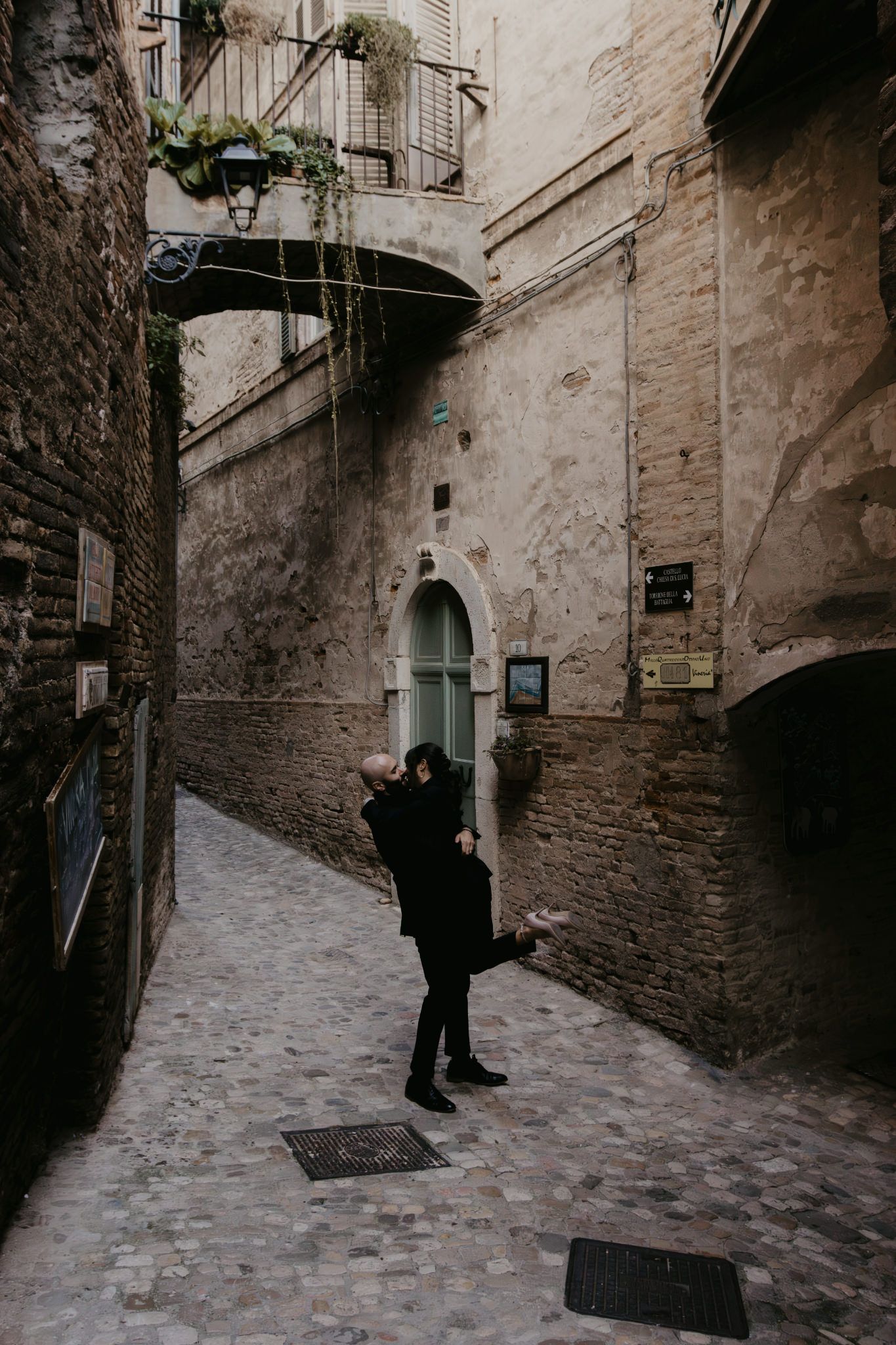 Couple embracing in a romantic cobblestone alley with ancient stone walls and arched doorway in an Italian town.