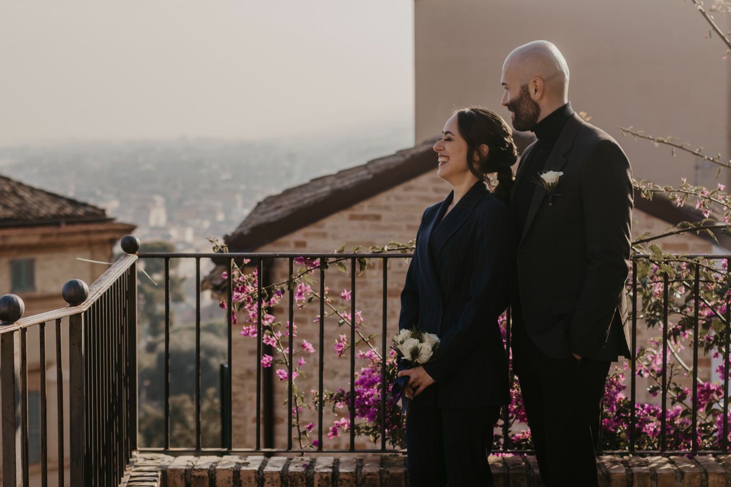 Couple in navy and black attire standing on a balcony with pink bougainvillea and city skyline views.