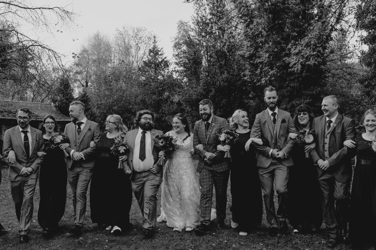 Black and white wedding party photo with bride, groom, and guests laughing outdoors among trees.