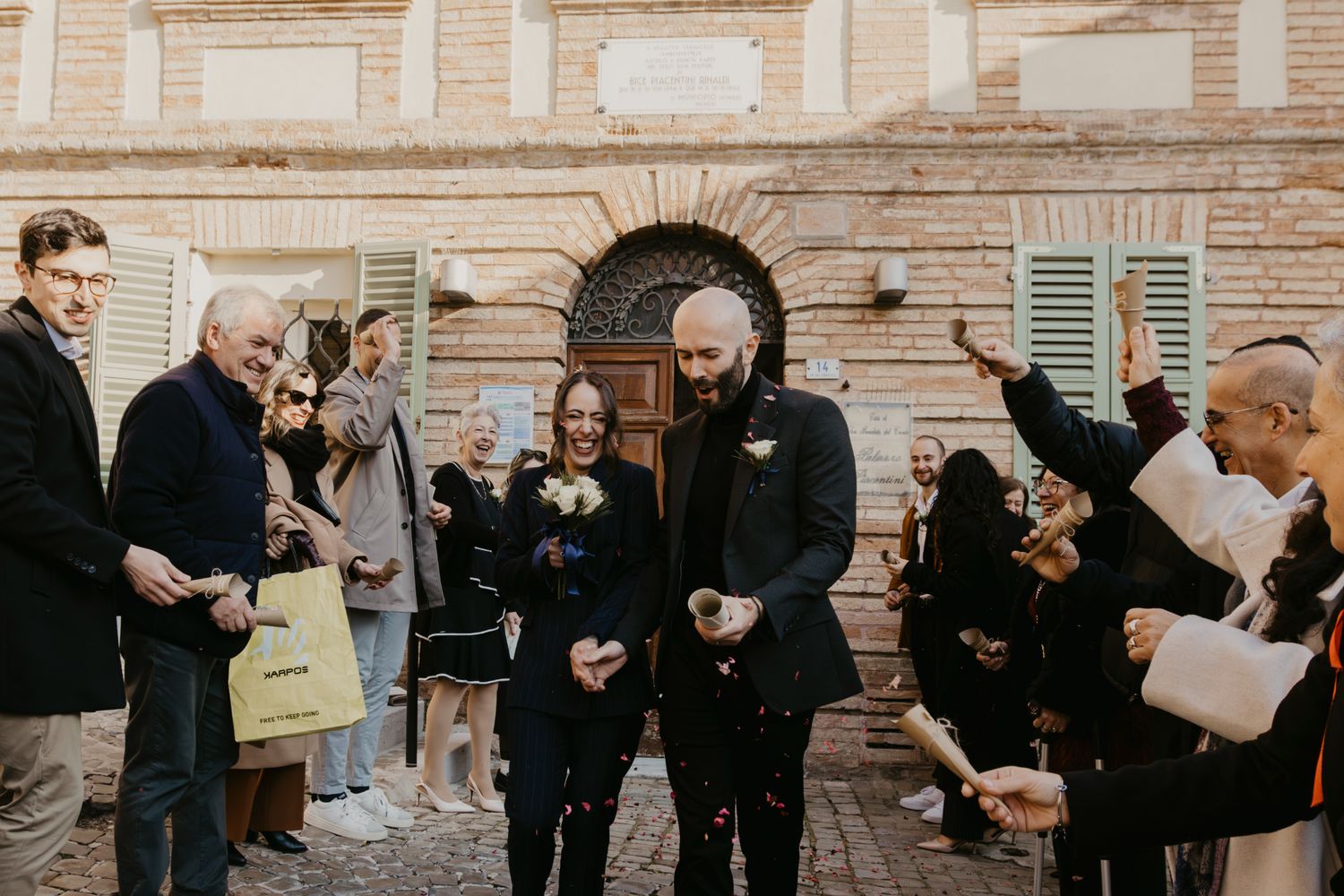 Joyful wedding couple celebrated with confetti by guests outside a rustic brick building.