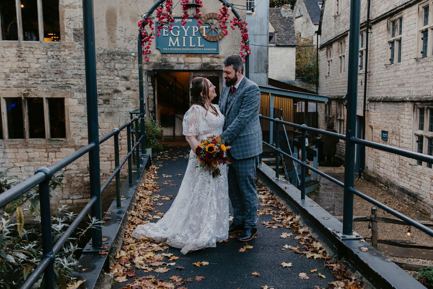 A couple in wedding attire stands on a rustic wooden bridge surrounded by autumn leaves and stone buildings.