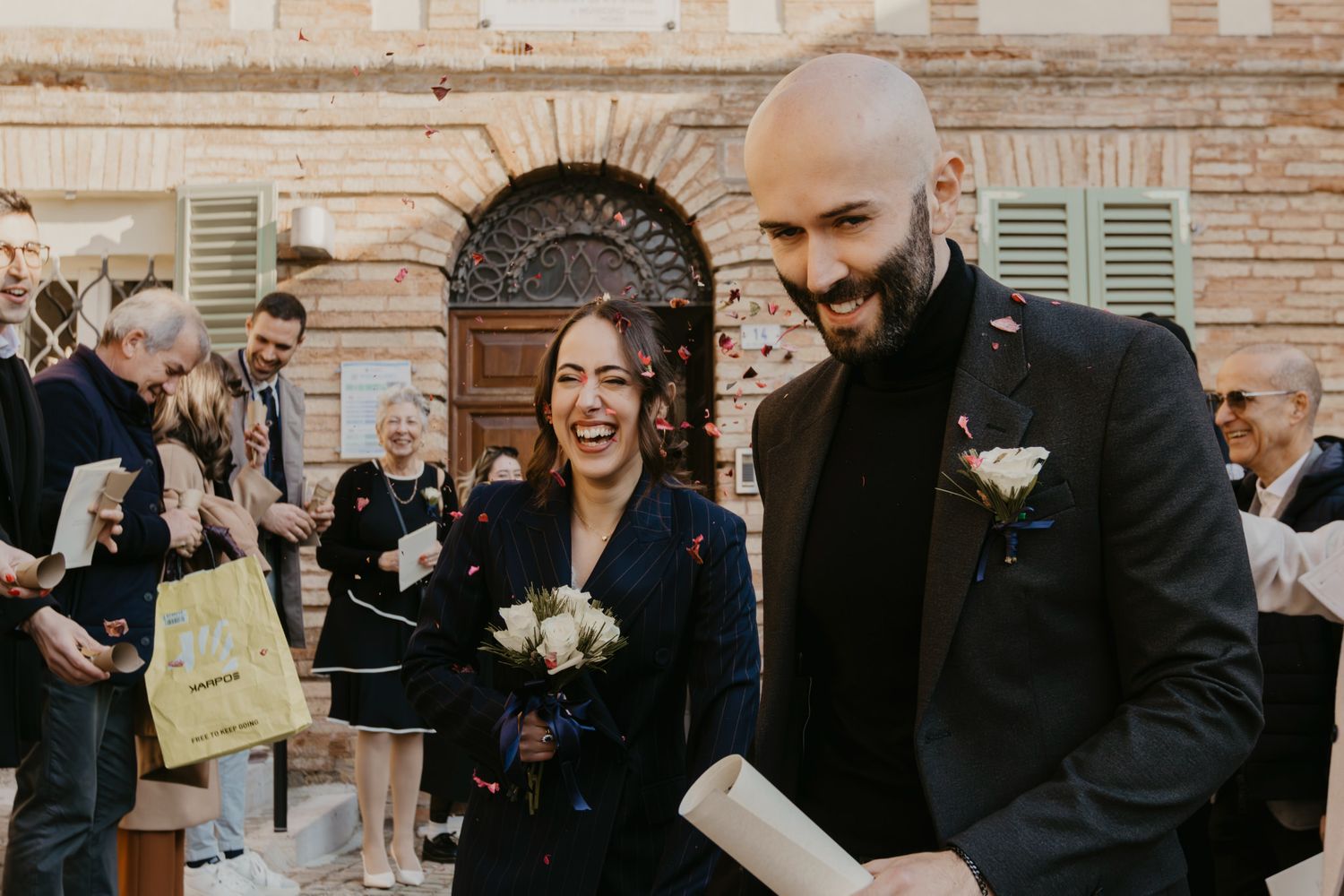 Joyful newlyweds laugh as guests toss confetti outside a rustic Italian building during their wedding celebration.