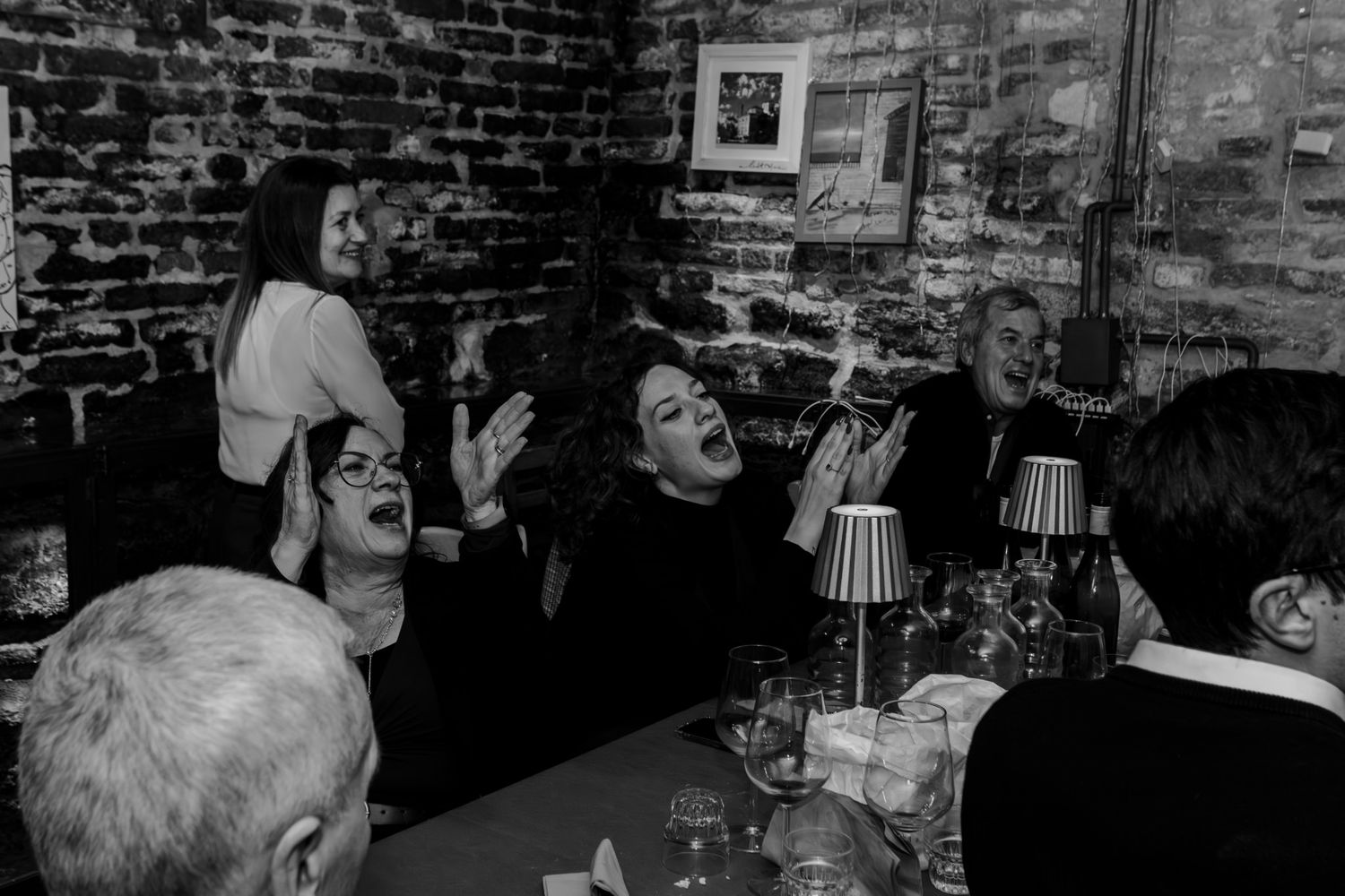 Black and white photo of animated women laughing and gesturing at a restaurant table with brick walls.