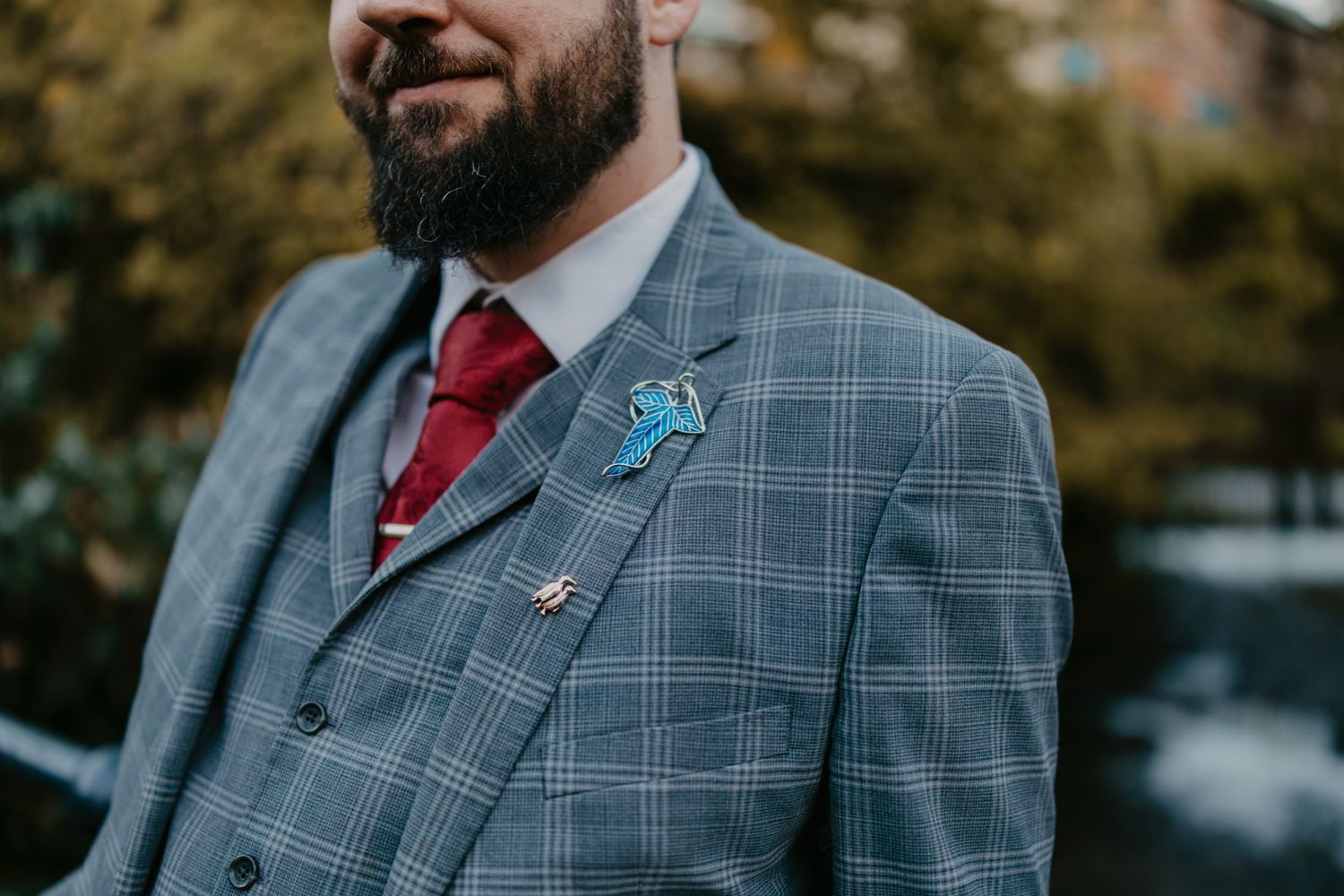 Bearded groom in grey plaid three-piece suit with red tie and blue floral boutonniere at wedding.
