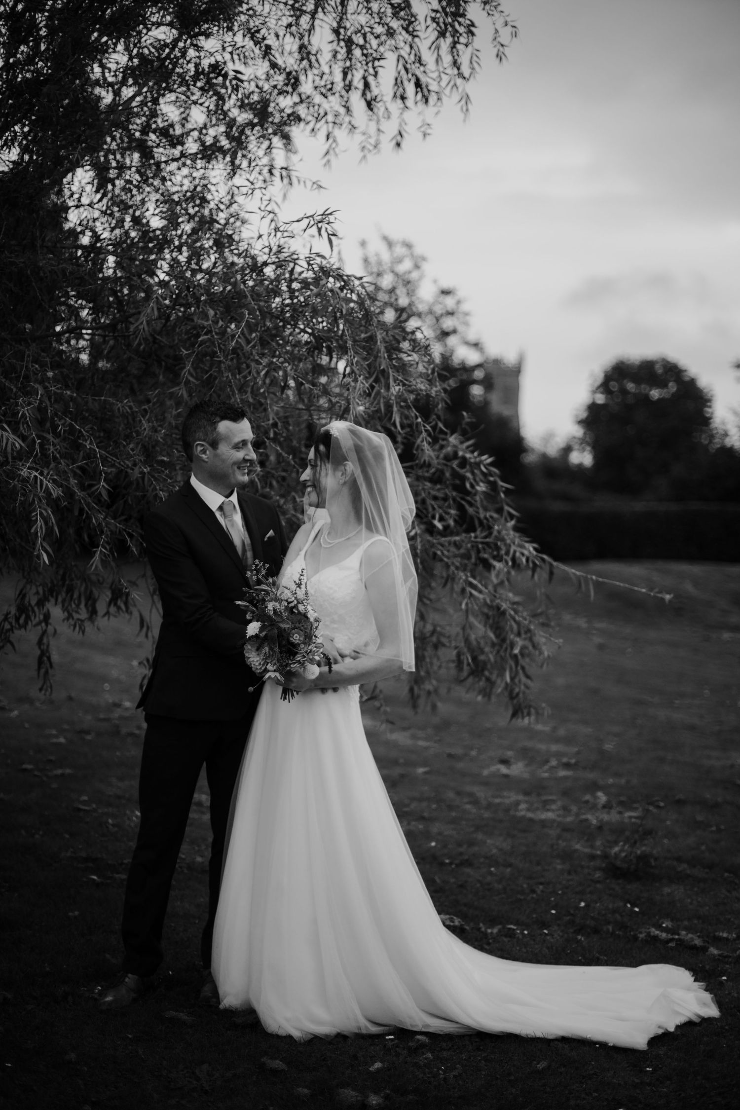 Black and white photo of a bride and groom standing under a willow tree, bride holding a bouquet, groom in a suit.
