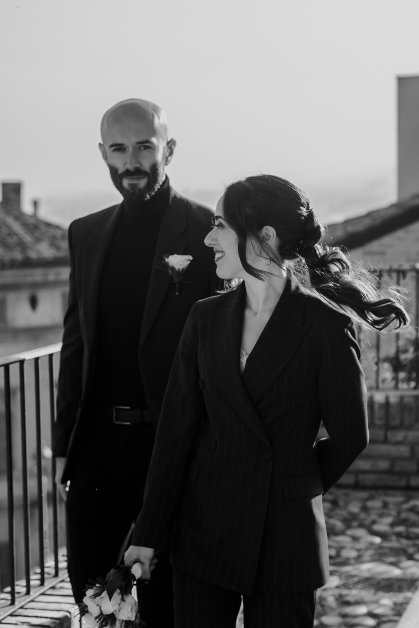 Black and white photo of a stylish couple in matching black suits on a balcony, woman smiling and holding flowers.
