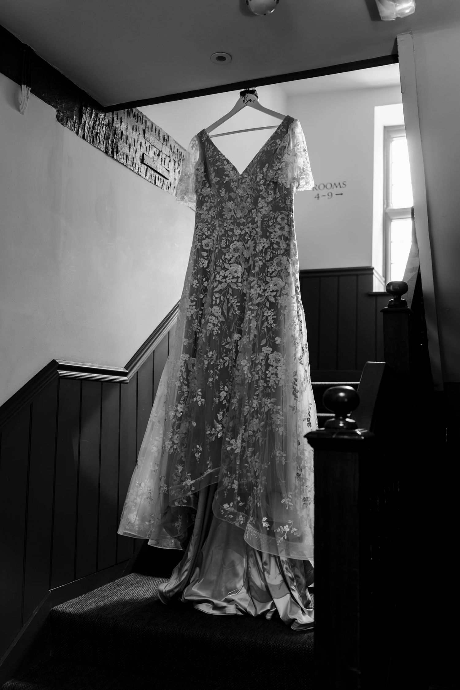 Black and white photo of a floral lace wedding dress hanging on a staircase banister near a window.
