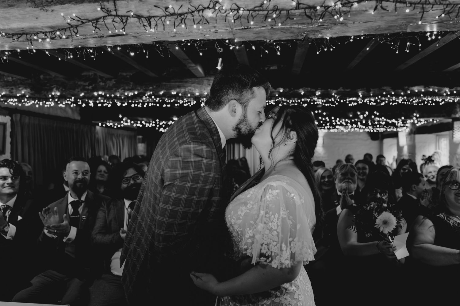 Black and white photo of a couple sharing their first kiss at a wedding ceremony, surrounded by guests in a fairy-lit venue.