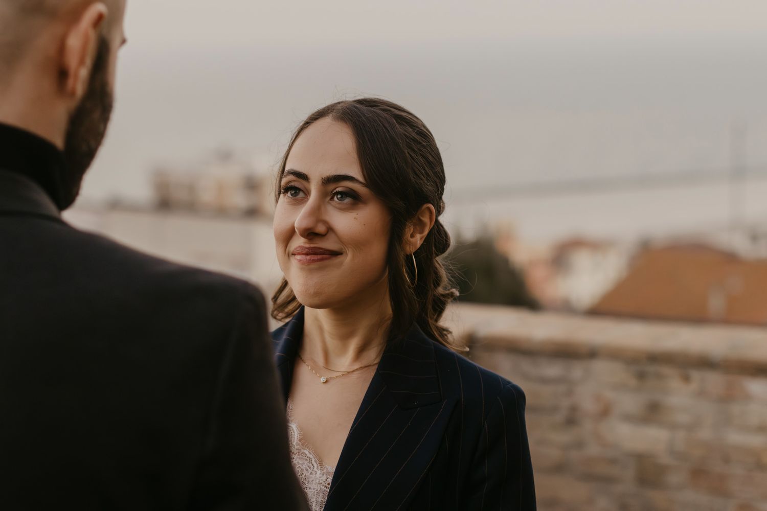 A bride smiles warmly at her groom during an outdoor wedding ceremony with a scenic coastal backdrop.
