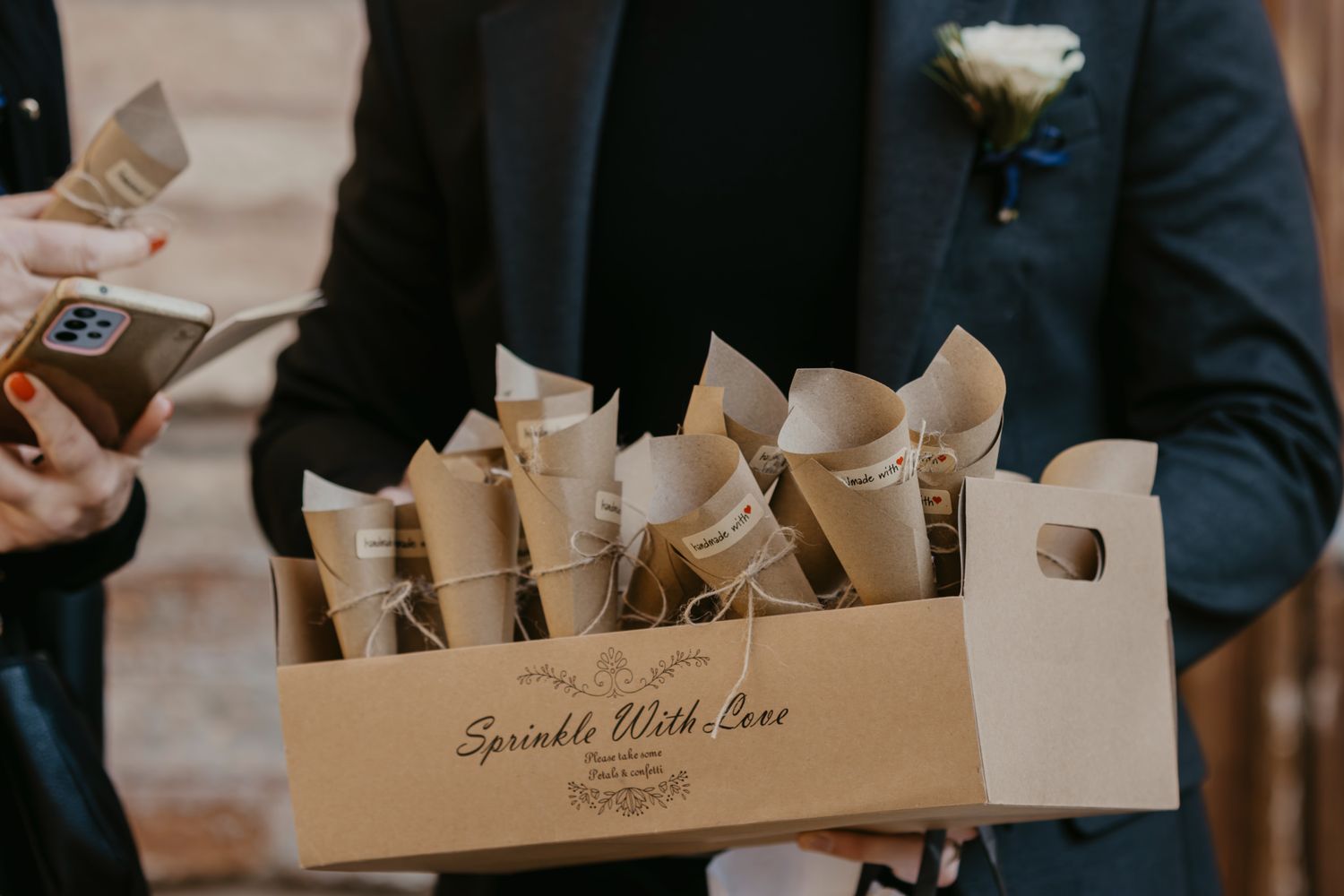 Groomsman holding a kraft box of paper cone confetti packets labeled Sprinkle With Love at a wedding.