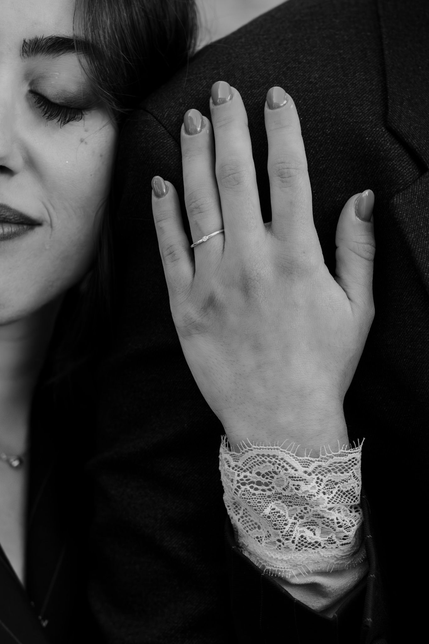 Black and white photo of a woman with closed eyes, hand resting on a dark jacket, wearing a delicate ring and lace cuff.