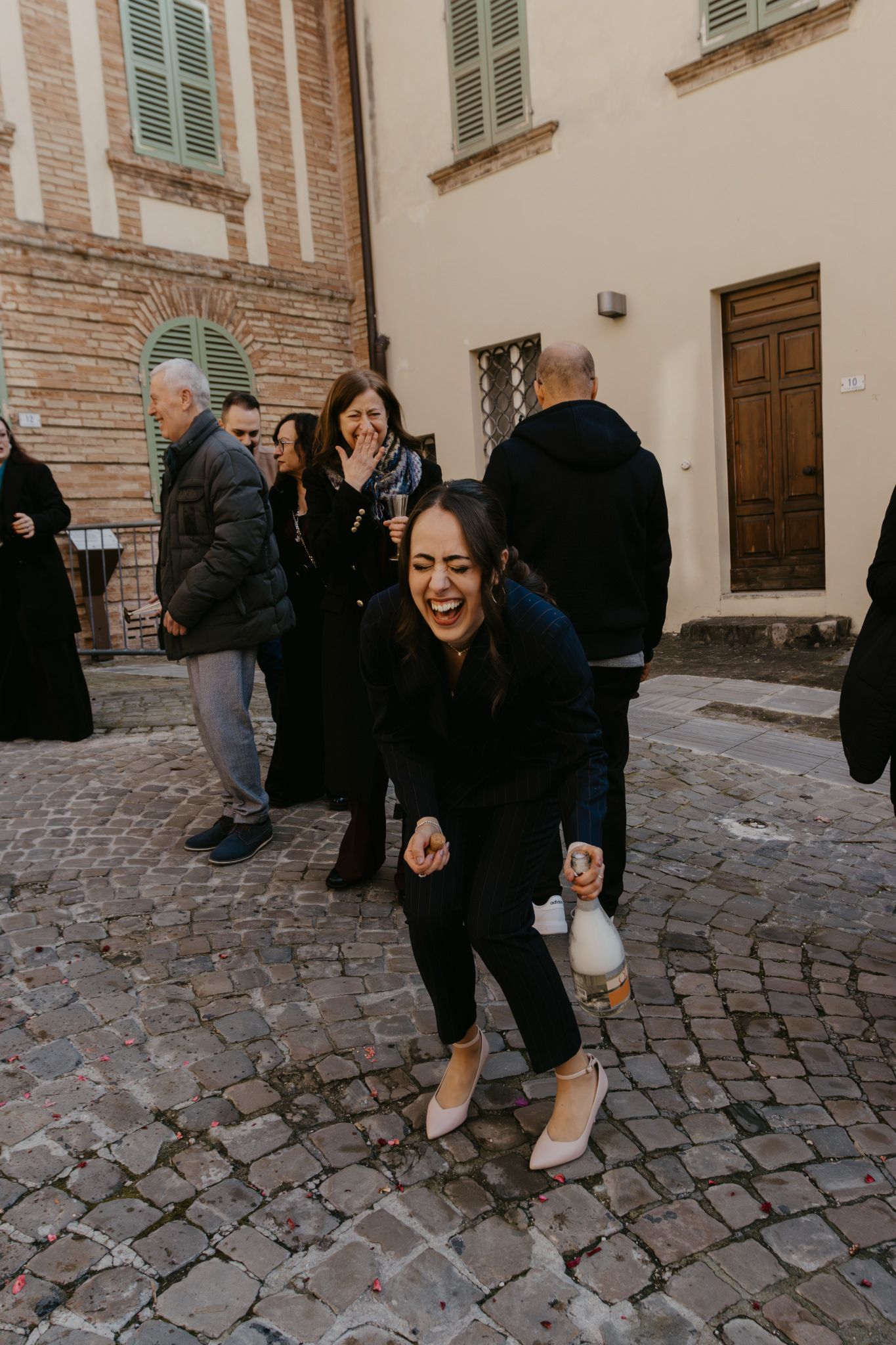 Woman laughing joyfully on cobblestone street in Italian town, holding bottle, surrounded by people.