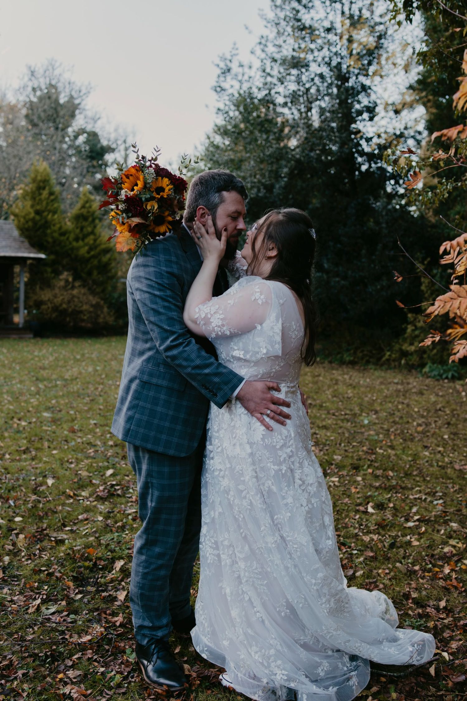Bride and groom share a romantic kiss outdoors in autumn, bride in lace gown holding fall bouquet.