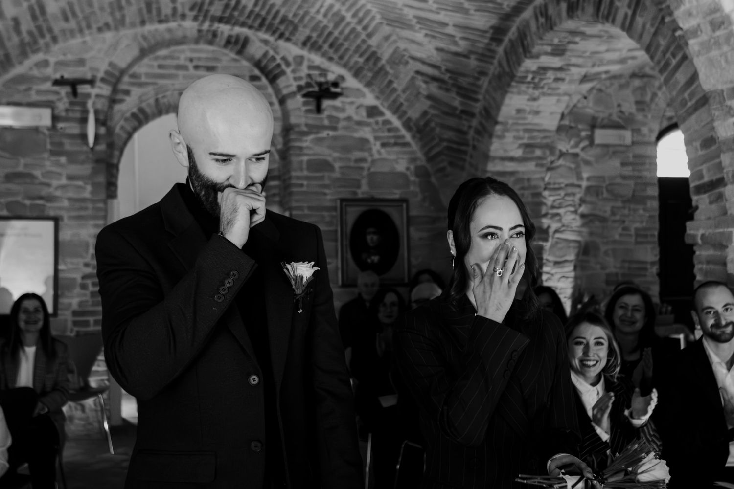 Black and white photo of emotional couple laughing at wedding ceremony in rustic brick arch venue.