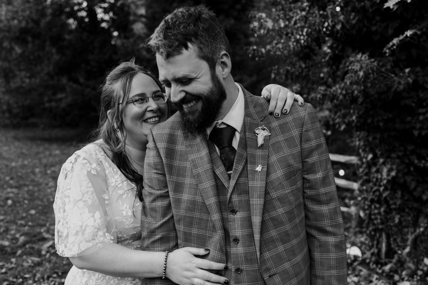 Black and white photo of a smiling bride and bearded groom in plaid suit sharing a joyful moment outdoors.