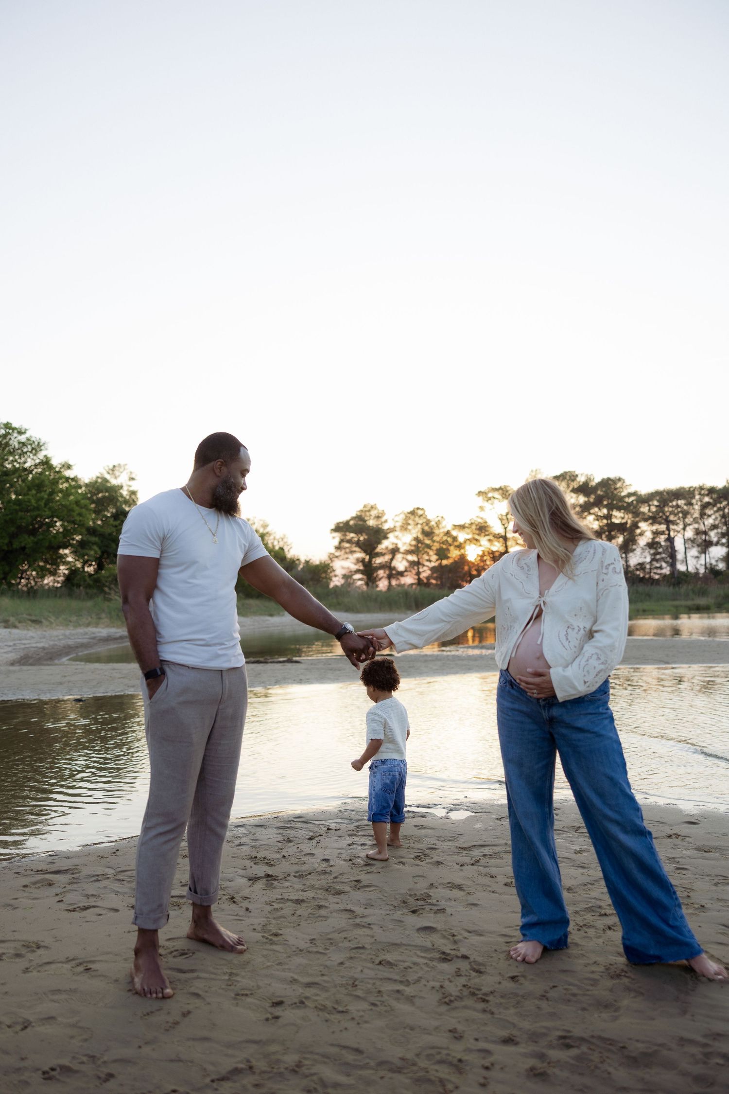 A pregnant woman and man hold hands with their toddler on a sandy beach at sunset.