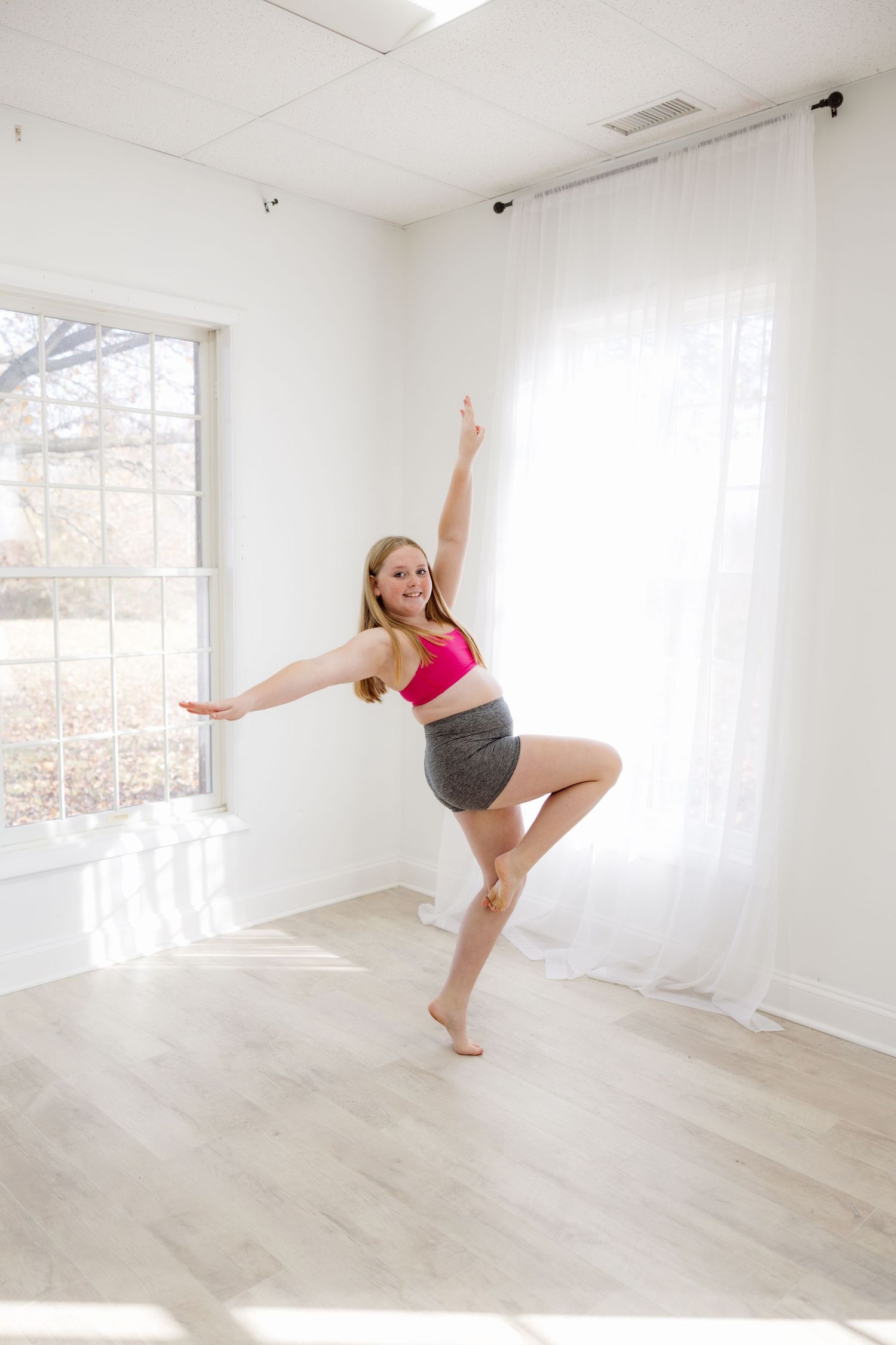 Dancer in pink performs graceful movements in bright white studio with natural light streaming through windows.