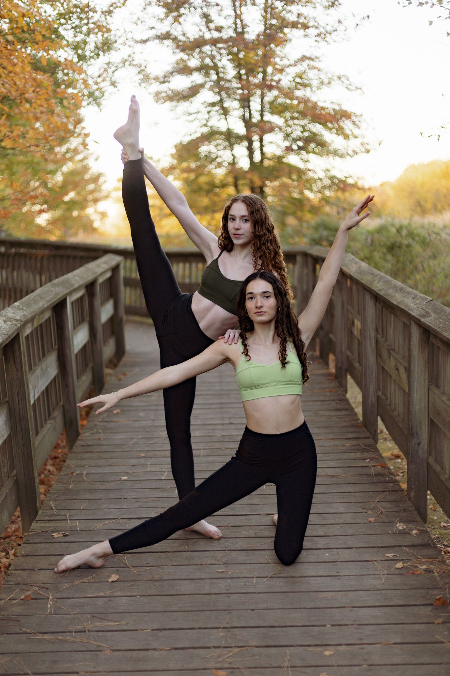 Two dancers perform synchronized leg extensions on wooden boardwalk during sunset.