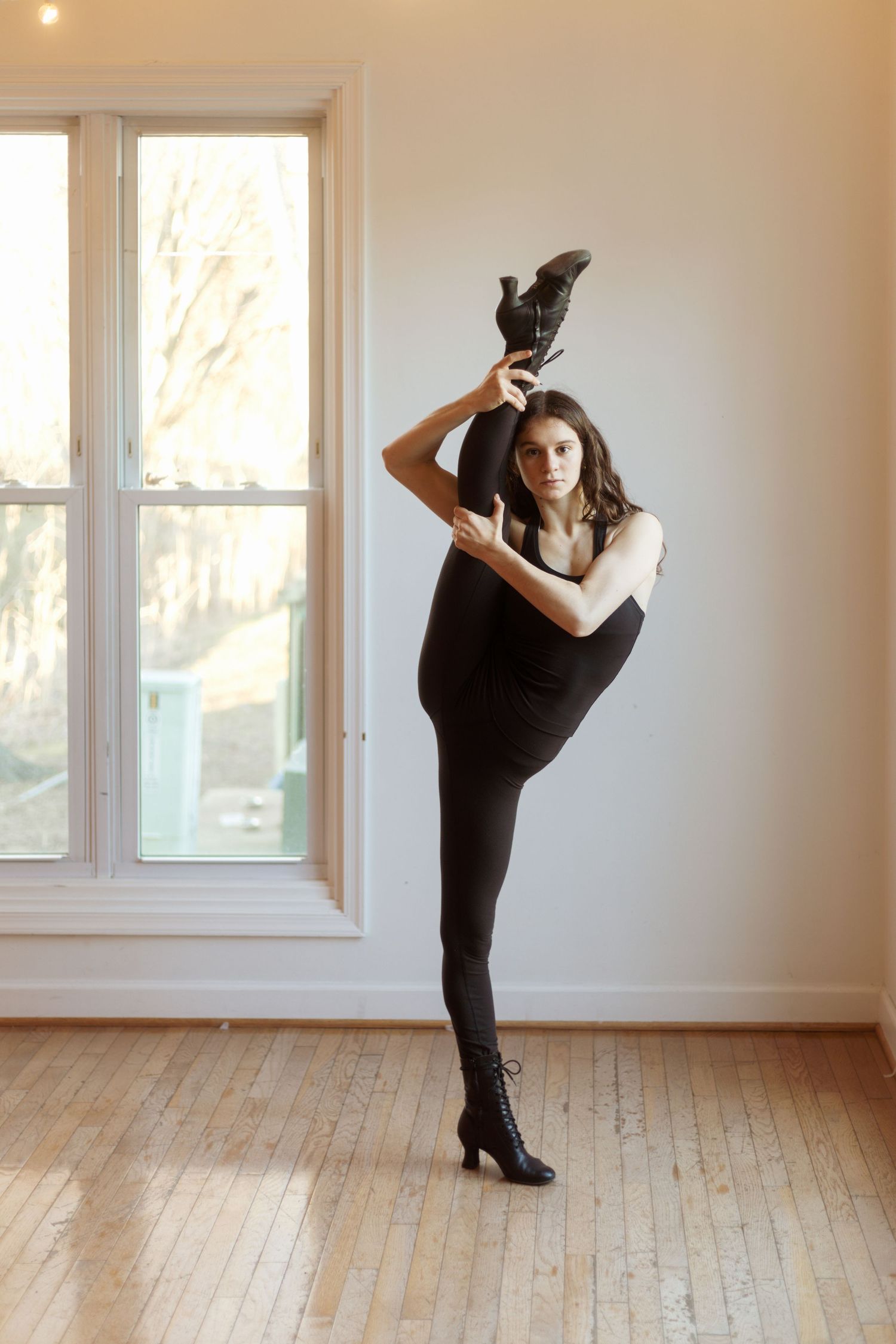 Athletic dancer performs high leg extension while wearing all black against bright windows.