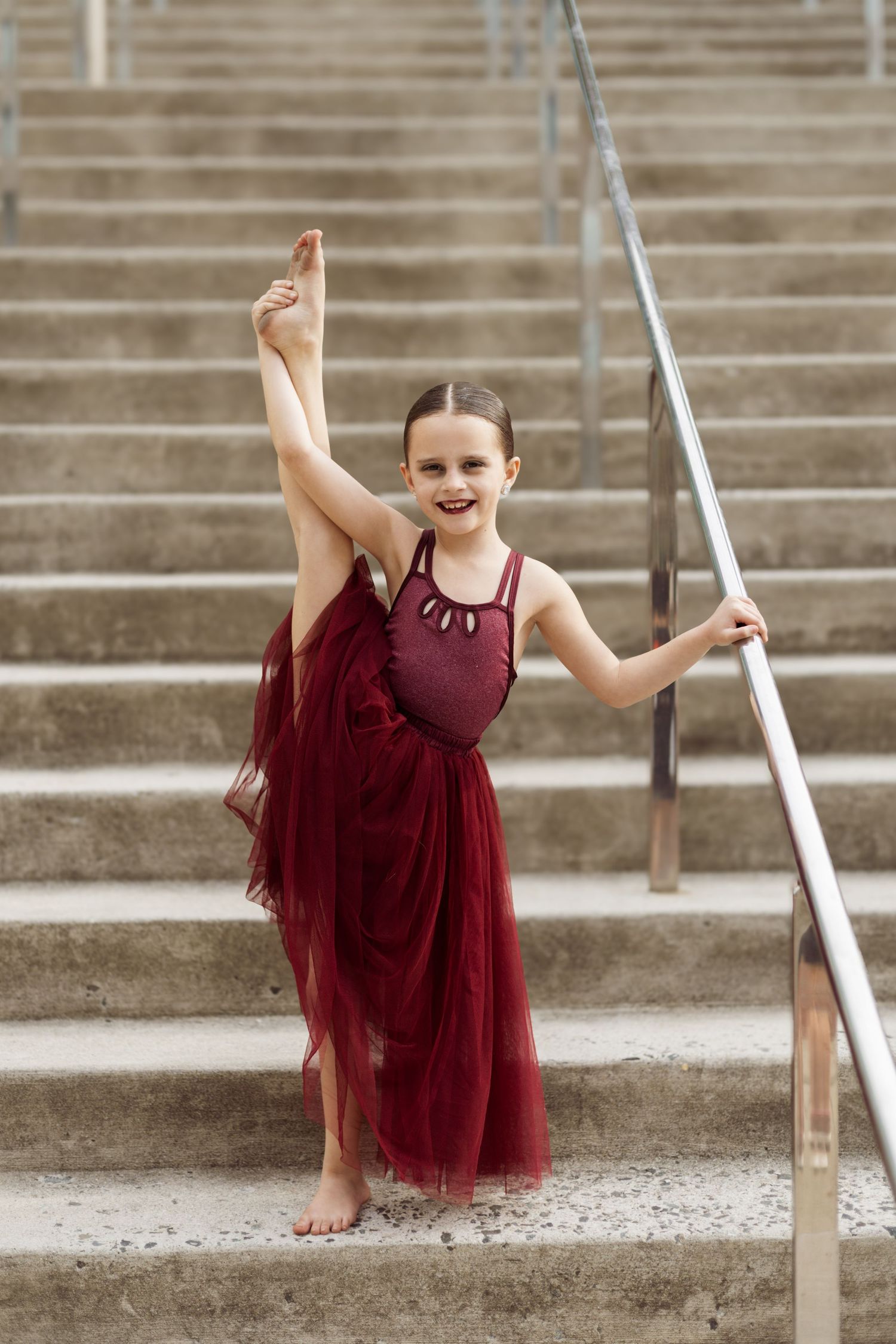 Young dancer in flowing burgundy tulle dress poses gracefully on concrete steps.