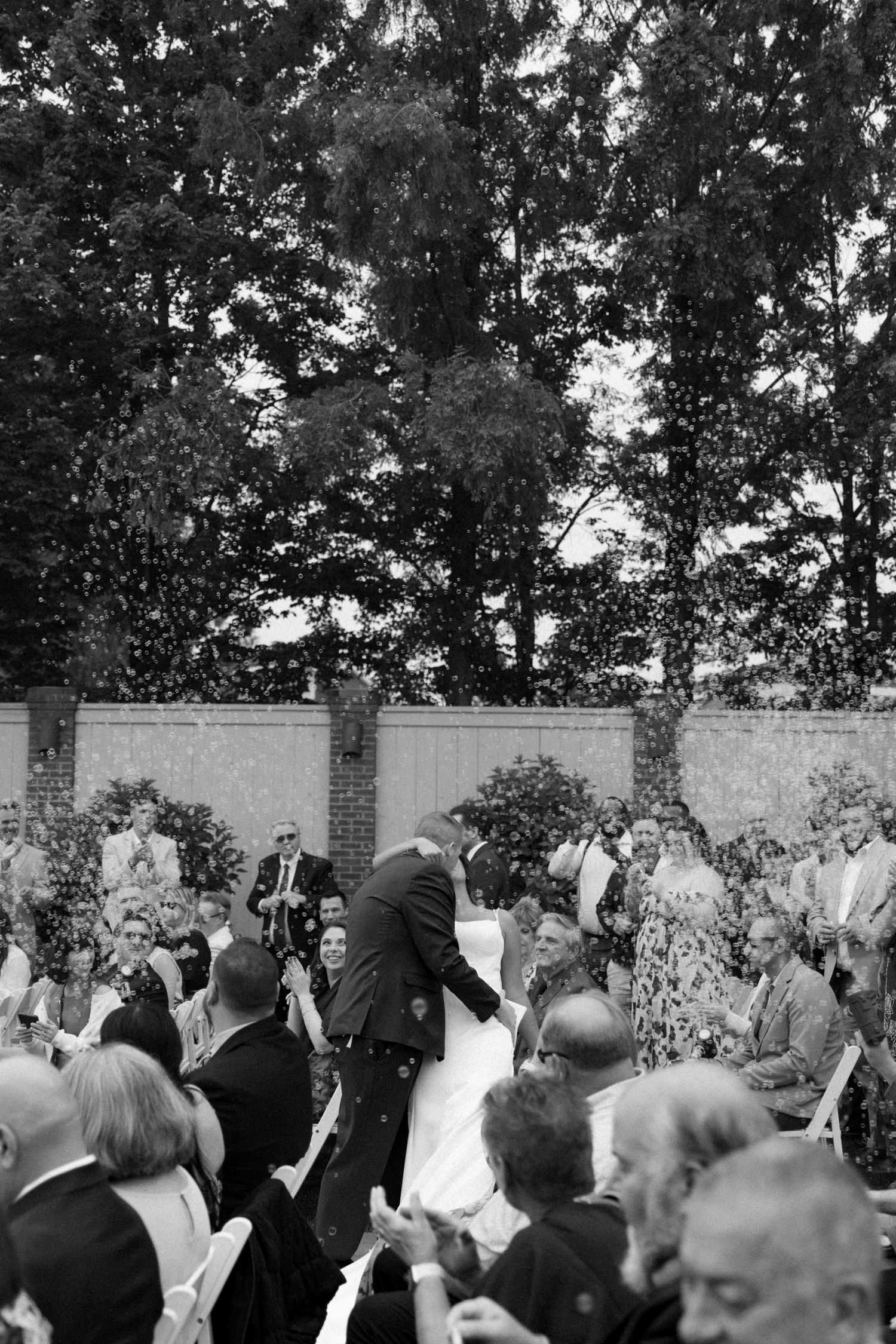 Black and white outdoor wedding ceremony with guests seated, couple at altar, confetti falling, surrounded by trees and brick wall.
