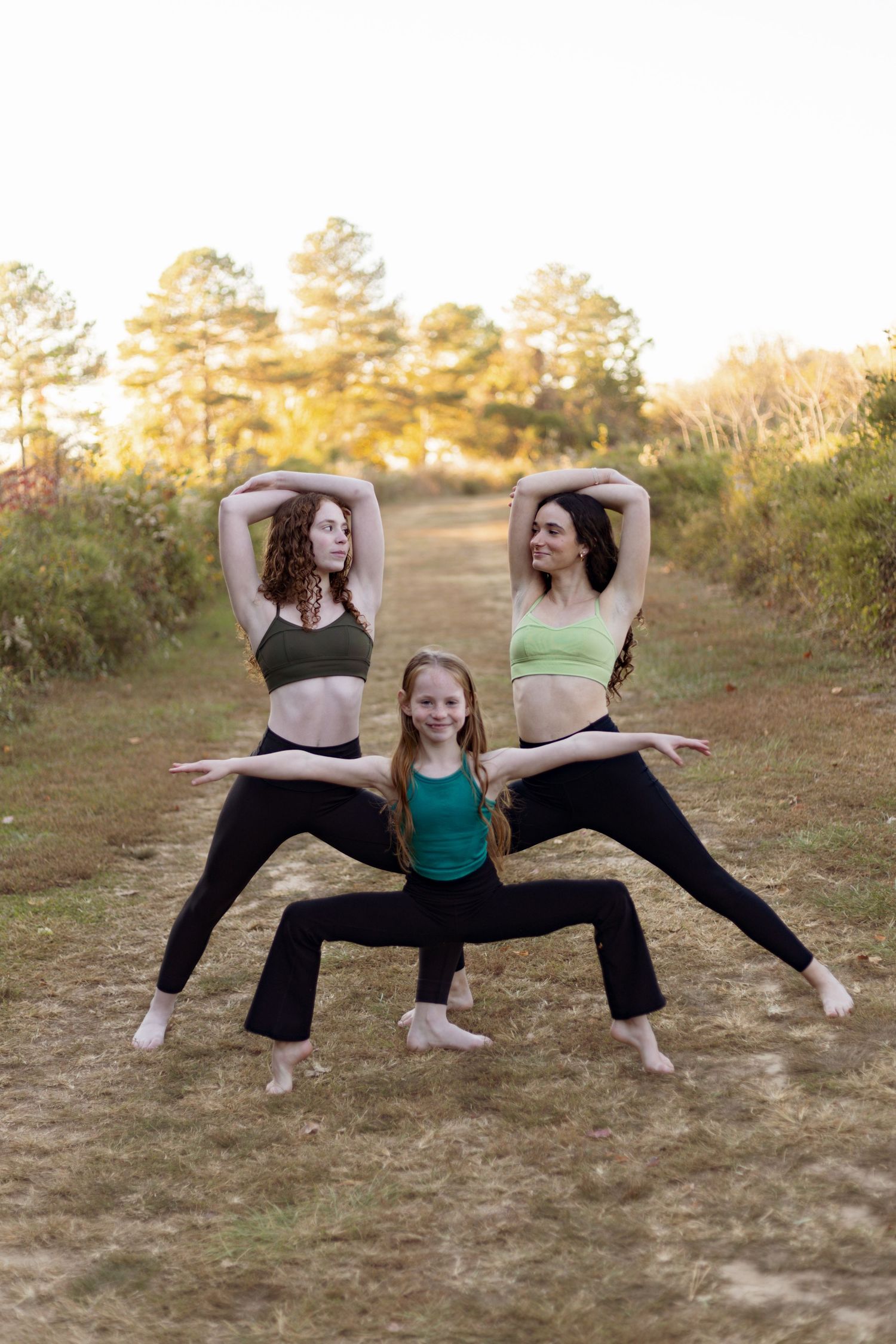 Three dancers perform synchronized poses outdoors with pine trees in background.