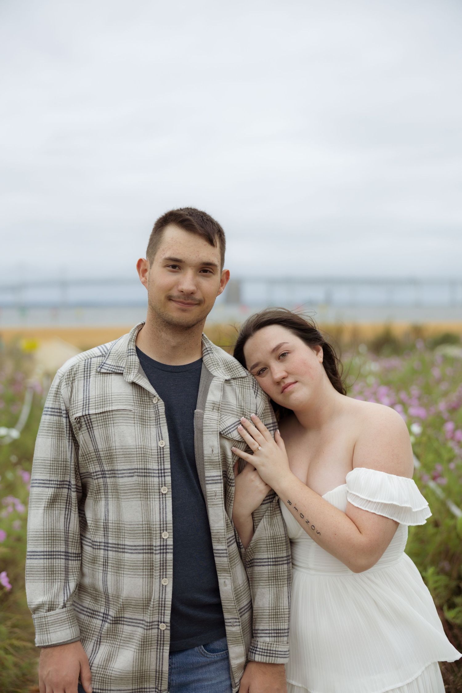A couple poses together in a field of wildflowers, with the person in white leaning against their partner in plaid.